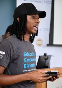 Young man wearing a hat and t-shit that reads "Educate, Empower, Elevate" speaks